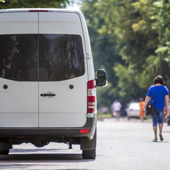 Back view of white passenger medium size commercial luxury minibus van parked n shadow of green tree on summer city street i with blurred silhouettes of pedestrians and cars under green trees.